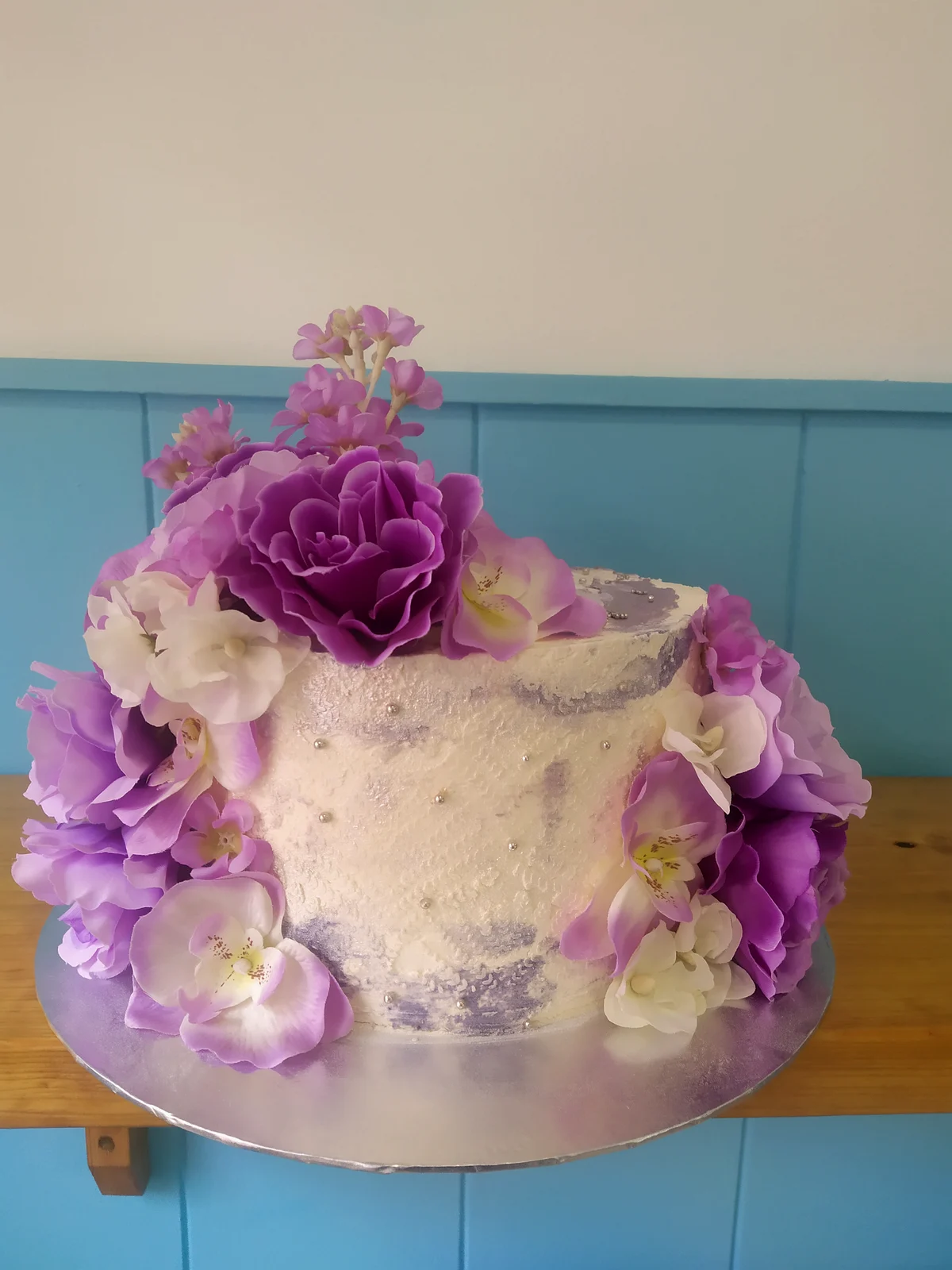 Tarta blanca rodeada de flores naturales moradas, lilas y blancas
